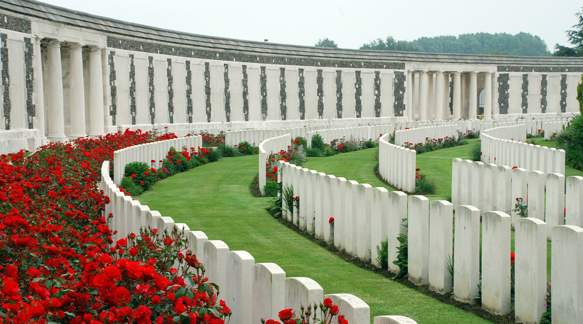 White stone headstones in long curved rows on green grass with read flowers and a larger white building behind